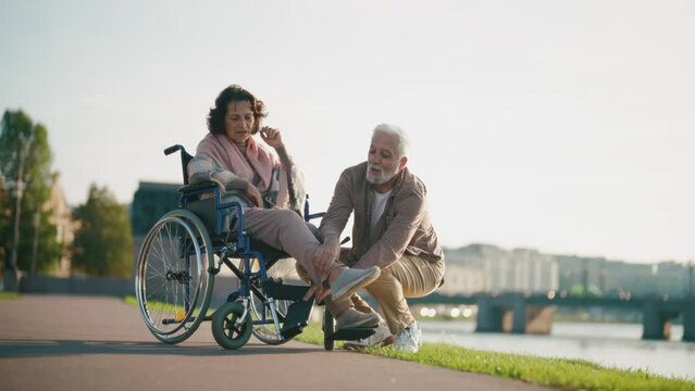 Wife in wheelchair makes couple. Senior happy family spending time in city park near pond taking photos on smartphone. Man hugging woman showing love care. Living with physical disability.