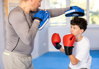 Sportive teenage boy training boxing kicks on punch mitts held by instructor in sports hall