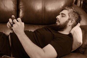 Happy guy using smartphone, enjoying leisure at home, relaxing on leather couch, overusing mobile phone, texting message, chatting online. Cellphone addict, social media obsess