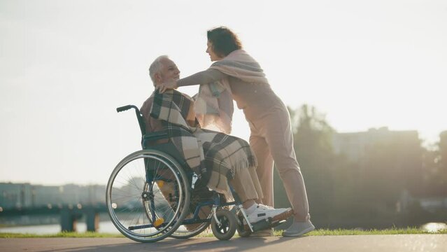 Wife helps husband to sit in wheelchair. Woman putting senior man legs on wheelchair footplates, walking together in city park. Caring female help male with special needs, physical disability concept.