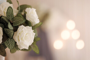 Close-up of white roses and green leaves against a backdrop of soft, blurred lights. Concept for wedding decorations or elegant events with ample copy space.