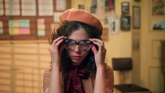 young woman in vintage clothes in yellow beret take off her retro glasses and looks tiredly at camera. emotionally burnt-out girl in office in the evening after working at computer or typewriter.