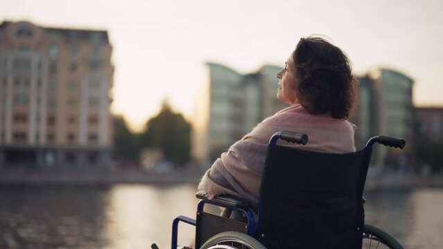 Woman in wheelchair walking in city. Senior mature smiling woman breathing air outdoor thinking looking around enjoying pastime in evening. Live with physical disability, special needs people concept.
