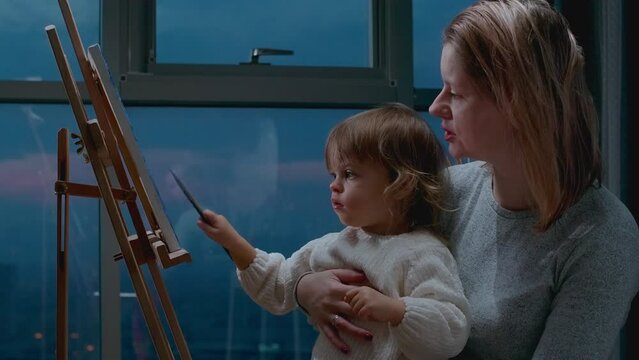 Mother teaching her little toddler how to draw a picture with oil paintings. Drawing together with momy, leisure activity with child at home.