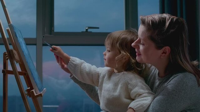 Mother teaching her little toddler how to draw a picture with oil paintings. Drawing together with momy, leisure activity with child at home.