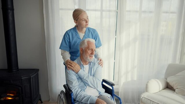 Nurse woman cares old man in wheelchair. Social medical worker pushing wheelchair with paralyzed senior male helping supporting him at home. Living with spine legs paralysis, special needs people.