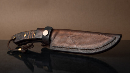 Ein handgeschmiedetes Messer mit einem Griff aus schwarzem Horn in einer ledernen Scheide auf schwarzem Glas, was das Messer spiegelt