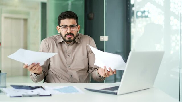 Confused businessman in glasses having difficulty with paper work sitting at workplace in business office. Puzzled entrepreneur looks at documents and cannot understand the problem looking at camera