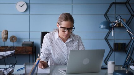 young woman in white shirt and glasses sits at table in office with laptop works multitasking. types on keyboard, makes notes, talks phone, looks concentrated and hard working under deadline pressure