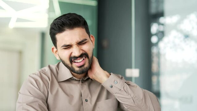 Tired bearded businessman suffering from neck pain while sitting at workplace in modern business office. Upset exhausted male employee in a shirt massages and rubs sore muscles, stretches. Close up