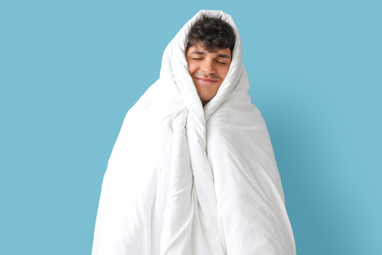 Handsome happy young man wrapped in soft blanket sleeping on blue background