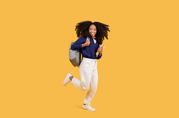 Fototapeta na wymiar Happy black student lady with backpack and copybooks, joyfully jumping on yellow background