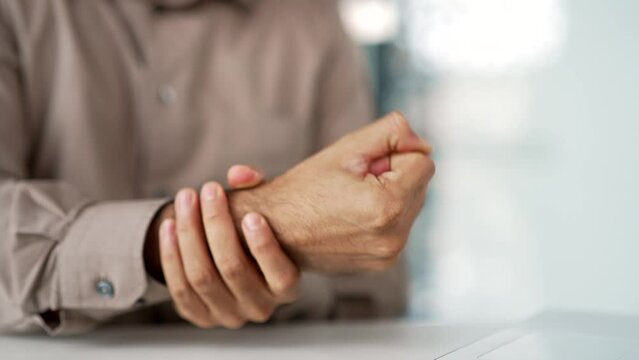 Close up of a man's hand massaging his wrist while sitting at a workplace at a desk in a business office. A male employee suffers from joint pain, has an injury, rubs a sore muscle, does an exercise
