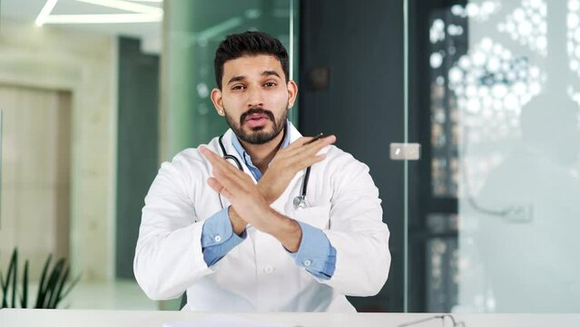 Serious doctor in white coat showing crossed arms, X sign, shaking finger no while sitting in modern hospital clinic. Medical worker physician demonstrates gesture of prohibition, denial, disagreement