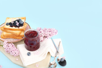 Board with jar of blueberry jam, toasts and flowers on blue background