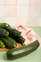 Board with many fresh zucchini on green wooden table near white tile wall