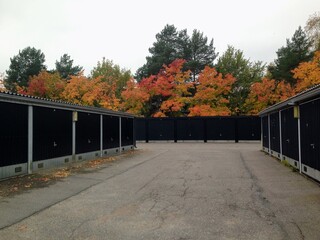 Beautiful autumn perspective view to the open space with cracked asphalt between low metal geometric shape warehouse garages with black doors and vibrant fall colours trees on the background in suburb