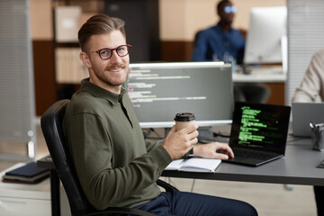 Medium long shot of smiling Caucasian male programmer in glasses looking at camera and holding...