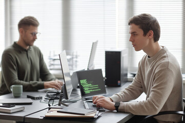Side view of young Caucasian male IT specialist looking at computer screen and typing on keyboard while sitting at desk with coworker in blurred background