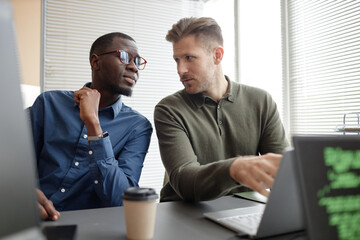 Medium shot of two diverse male programmers talking while sitting at desk in IT company office