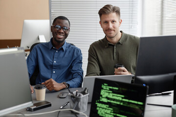 Portrait of cheerful African American IT specialist in glasses and Caucasian male coworker holding takeaway coffee cup looking at camera while sitting at desk
