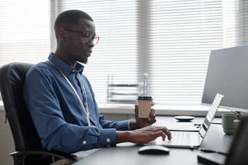 Side view of Black man in glasses typing on laptop and holding takeaway coffee cup while sitting at office desk in IT company