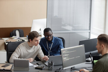 Wide angle shot of African American IT male mentor pointing out inaccuracies in code of young Caucasian male programmer while sitting with laptop at desk - obrazy, fototapety, plakaty