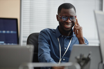 Waist up shot of cheerful Black man wearing glasses and blue shirt sitting in IT office while talking to customer using smartphone