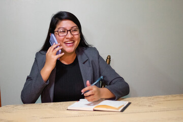 Mujer caucásica con chaqueta gris sentada cerca de la mesa hablando con el teléfono y sonriendo