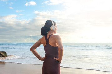 fit, sporty woman standing by the ocean with hands on her hips and wearing headphones