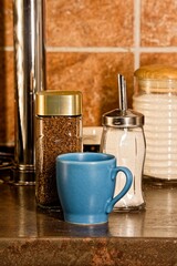 A blue cup, a jar of coffee and a sugar bowl stand on a table in the kitchen, a vertical photo with warm natural colors, a brown background