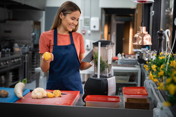 Smiling motivated young woman preparing healthy smoothie or vegetable side dish in restaurant kitchen using food blender mixer processor looking at digital tablet fintech device.