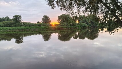 On an early summer morning, the sun rises above the trees on the far side of the river and colors the sky with delicate hues. The trees and cloudy sky are reflected in the water