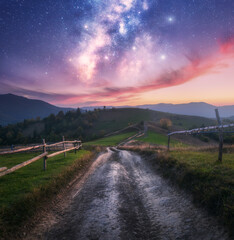 Milky Way over the rural road, wooden fence, green hills in mountain valley at summer night. Space. Landscape with country road, meadows, purple sky with stars at dusk. Carpathians, Ukraine. Nature