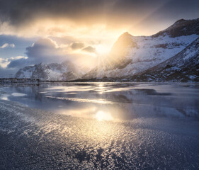 Beautiful snowy mountains and colorful sky with clouds and golden sunlight at sunrise in winter in Lofoten islands, Norway. Landscape with rocks in snow, frozen sea coast, reflection in water. Nature