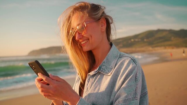 Smiling young woman with blond hair using smartphone on beach. Beautiful female in eyeglasses and denim shirt watching video on mobile phone. Lady enjoys her summer vacation on ocean shore.
