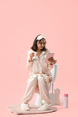 Young woman in pajamas with coffee using tablet computer on toilet bowl against pink background