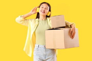 Young Asian woman in headphones with parcels on yellow background