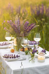 Romantic table decor for a loving couple on the blooming meadow with purple lupines. Two glasses of wine, flowers in a vase, silverware, fruits, wooden furniture and picnic basket. Sunset, golden hour