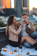 Surprise date on rooftop with urban cityscape and skyscrapers on background. Happy young loving couple drinking wine having romantic candlelit dinner celebrating anniversary or Valentines Day