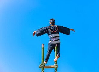 A Japanese hikeshi firefighter standing on one leg at the top of a tall bamboo ladder performing acrobatics during the traditional Hashigo-nori show of Suga shrine against the blue sky of Tokyo.