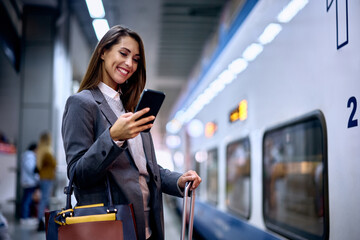 Happy woman texting on smart phone at train station. - obrazy, fototapety, plakaty