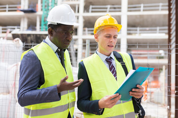 Two civil engineers working at the construction site are discussing the construction plan, holding documents in their hands and carefully studying them
