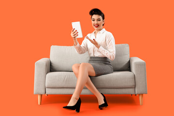Young pin-up businesswoman with tablet computer sitting on sofa against orange background
