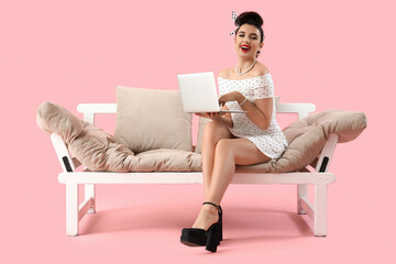 Young pin-up woman using laptop on sofa against pink background