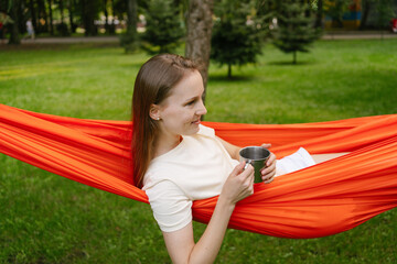 A young woman drinks tea from a metal mug while lying on an orange hammock hanging from a tree. The...