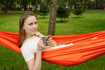 Young woman relaxing in an orange hammock in the trees with a metal cup of tea. The concept of...