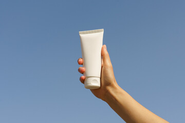 A female hand holds a white mockup of a blank tube of cream on a blue sky background. Concept of...