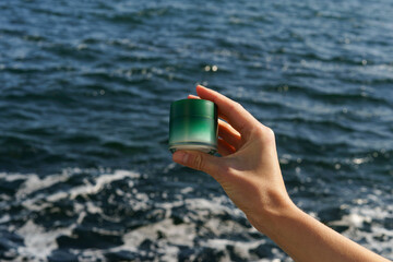 Female hand holding green face cream jar mockup on blue sea water background. The concept of natural beauty cosmetics