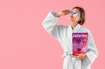 Beautiful young woman in bathrobe and with magazine on pink background